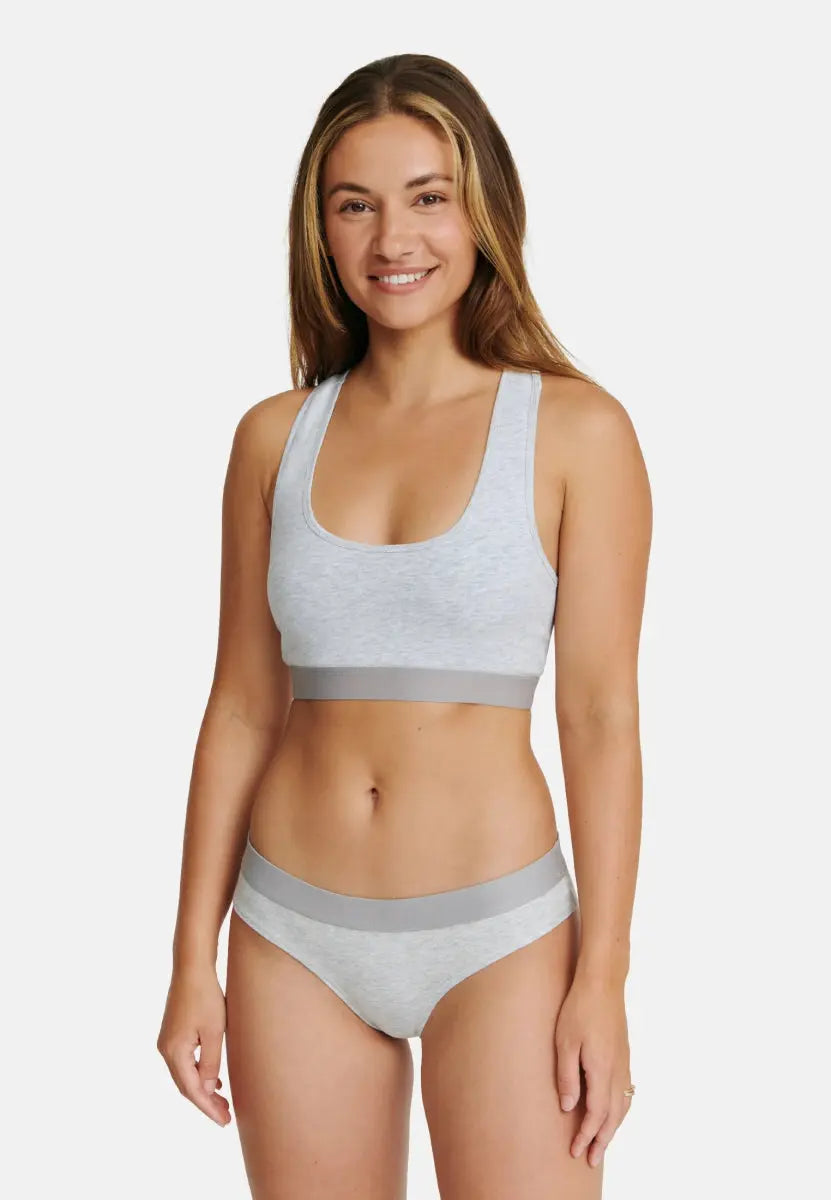 Woman in light gray organic cotton thong and matching sports bra, smiling against a white backdrop.