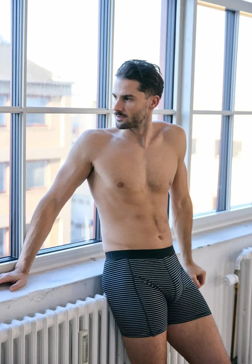 Man in striped black and white trunks, leaning against a window, looking away.