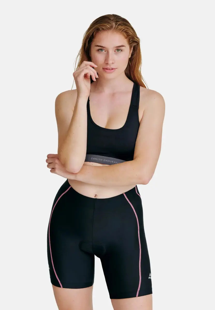 Woman in black padded bike shorts with pink trim, matching sports bra, standing confidently.