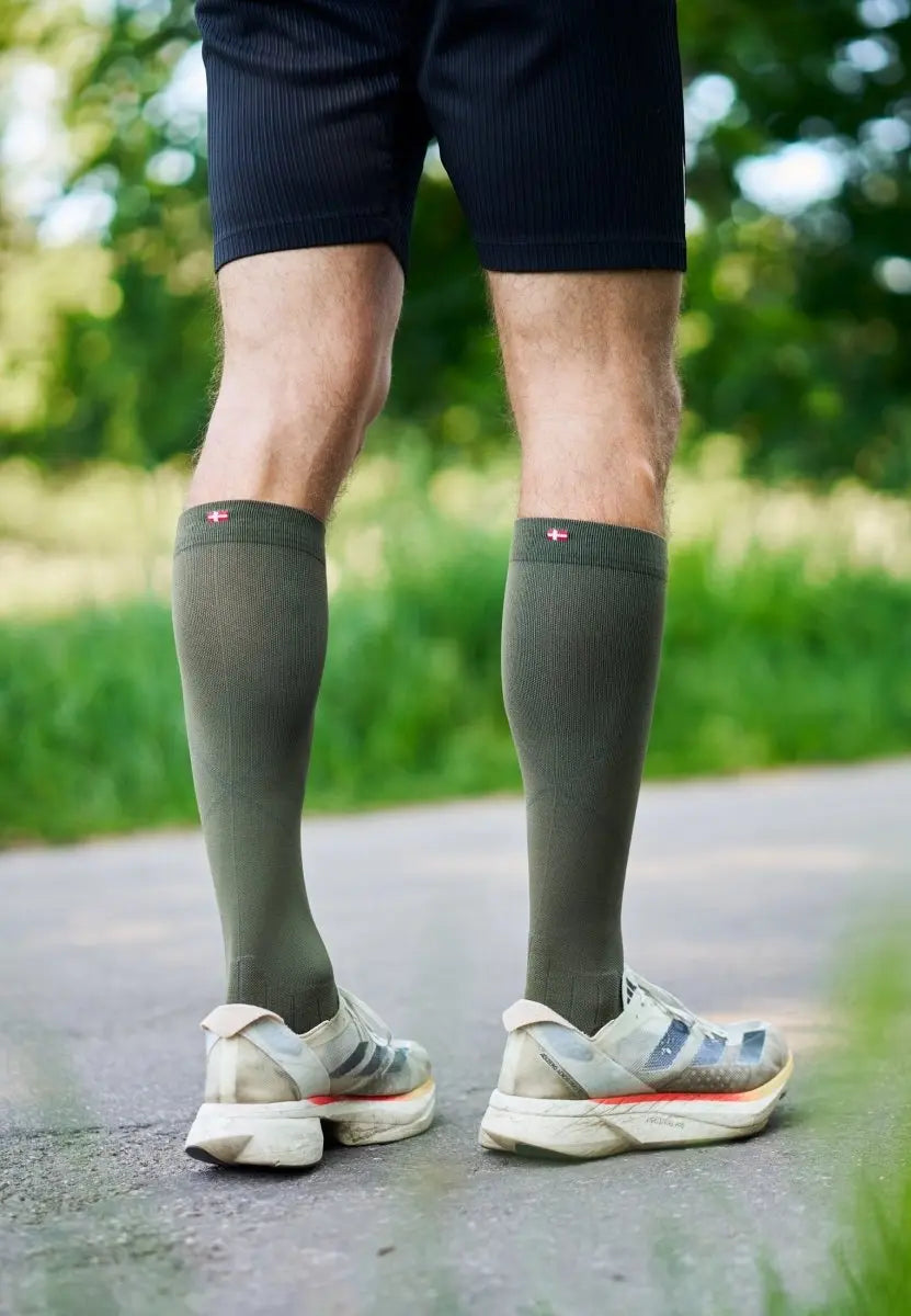 Person wearing olive green compression socks and sneakers on a path, with greenery in the background.