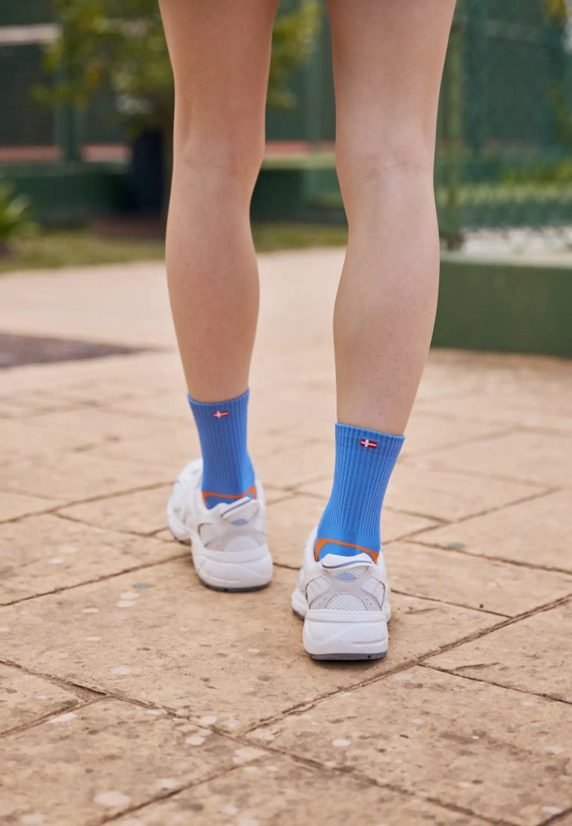 Blue sports socks with flag design, worn with white sneakers on a sunny paved path.
