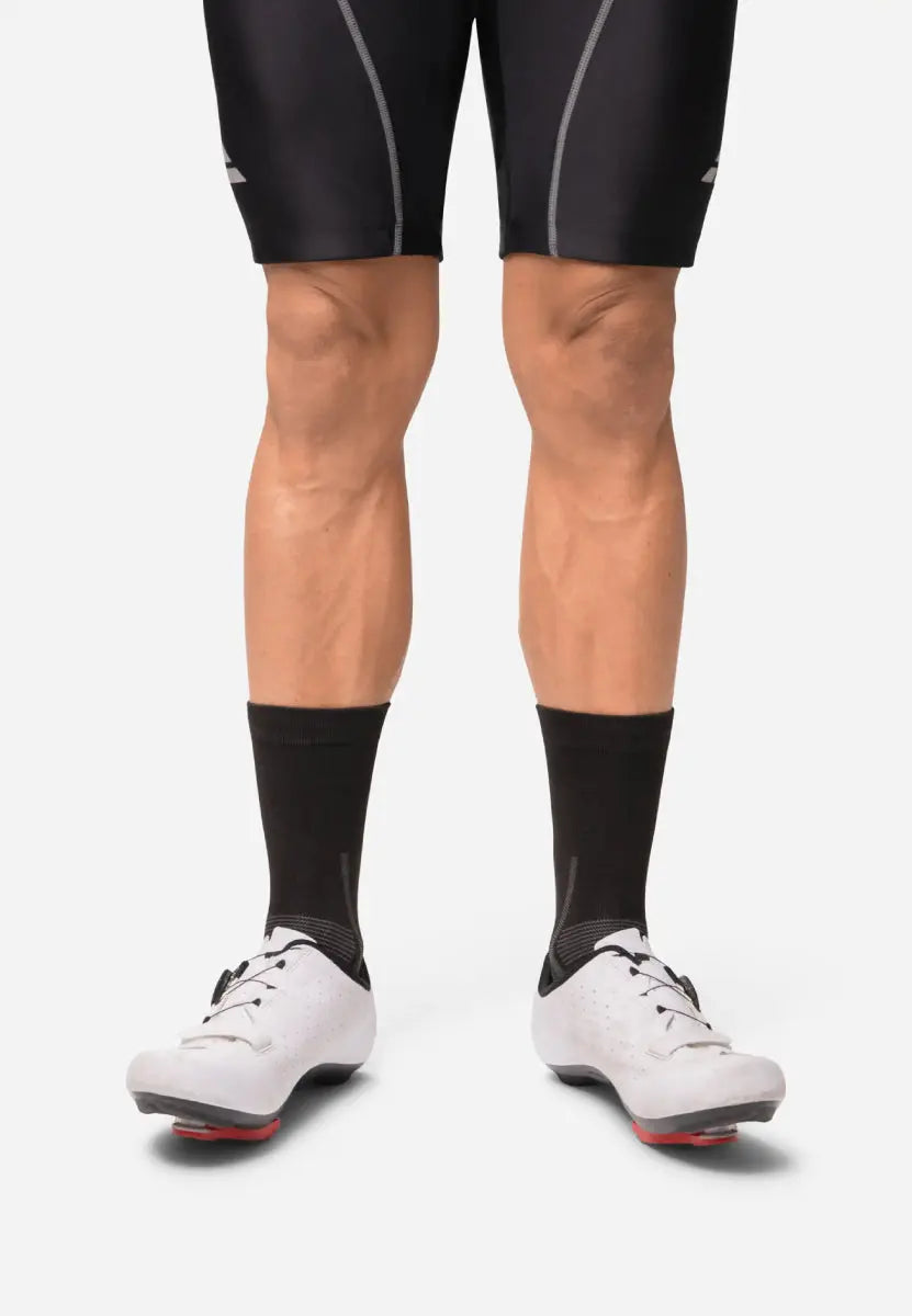 Person wearing black cycling socks and white cycling shoes, standing against a plain background.