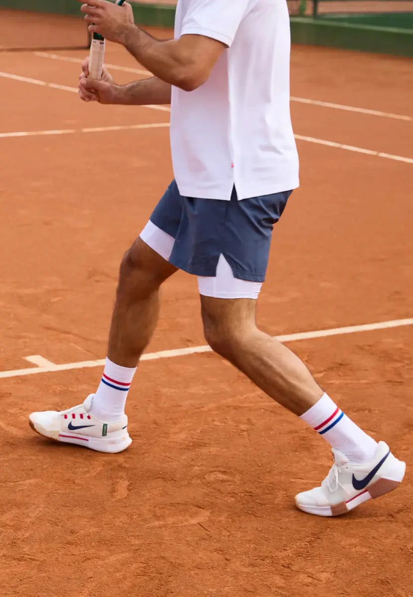 Person in tennis stance on clay court, wearing striped socks, white sneakers, blue shorts, white shirt.