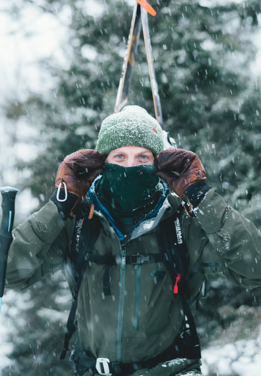 Person in winter gear with green beanie and neck gaiter, snowy background, holding ski poles.