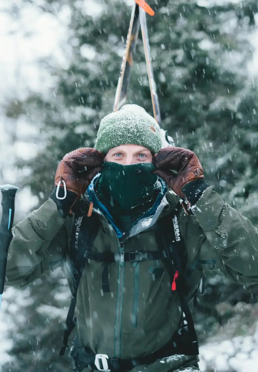 Person in winter gear with green beanie and neck gaiter, snowy background, holding ski poles.