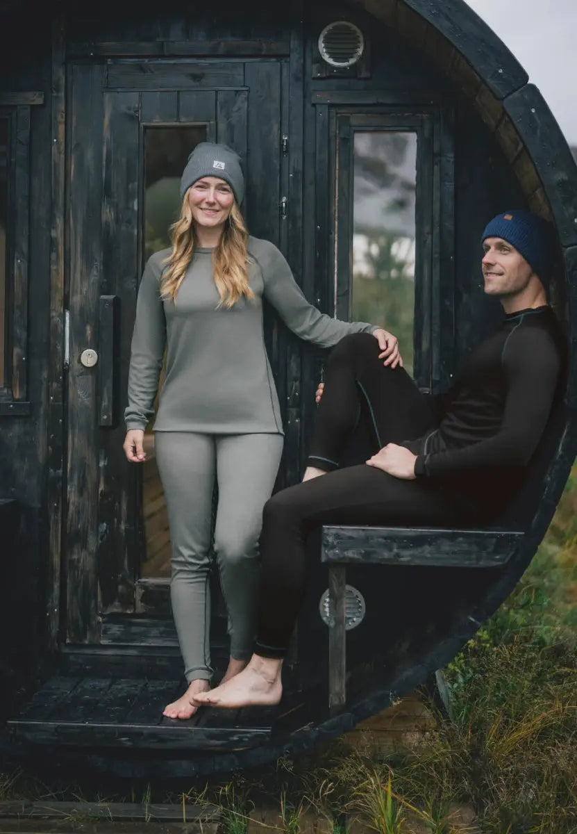 Two people in thermal underwear; one stands, the other sits, both smiling outside a wooden cabin.