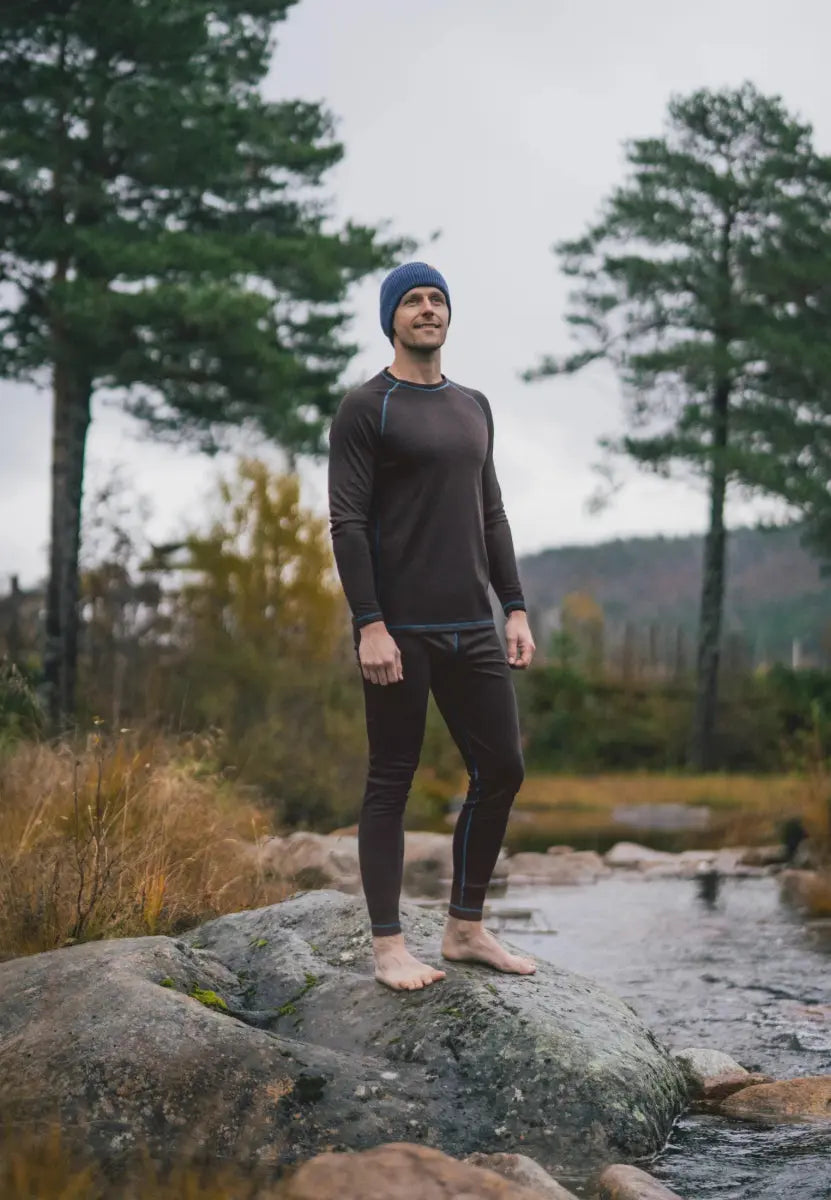 Person wearing thermal underwear and beanie stands on a rock by a stream, surrounded by trees.