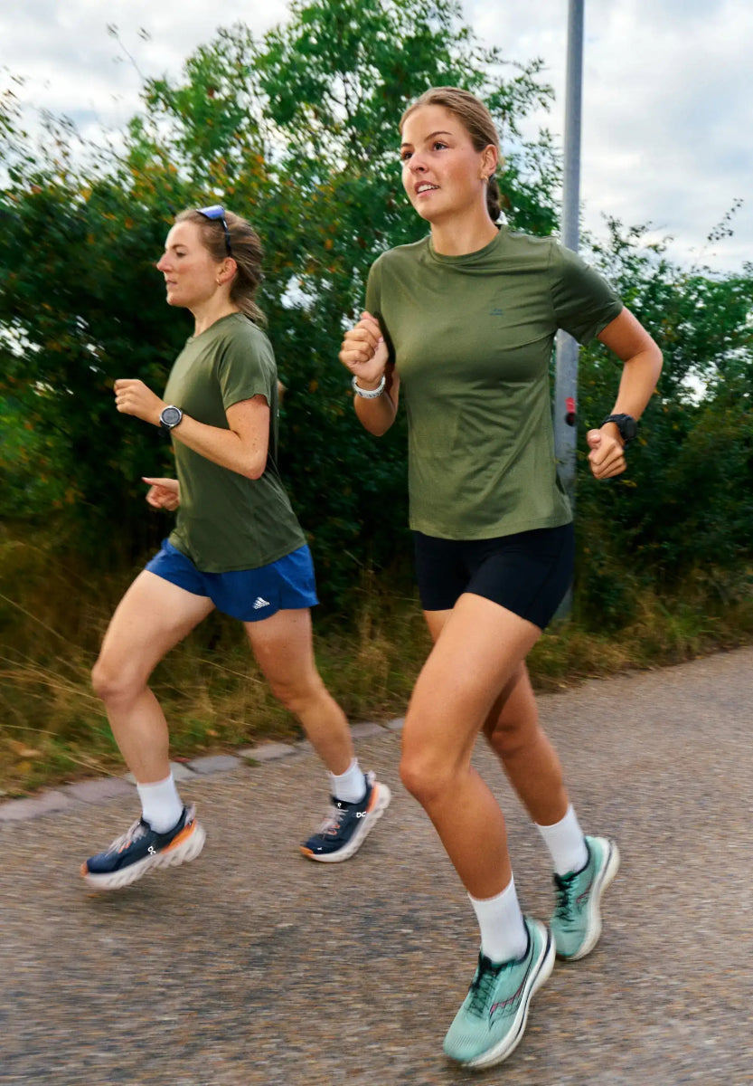 Two women jogging in green t-shirts and shorts on a path surrounded by greenery.