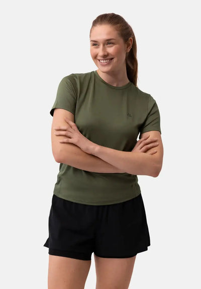 Woman in a green t-shirt and black shorts, smiling with arms crossed against a plain background.