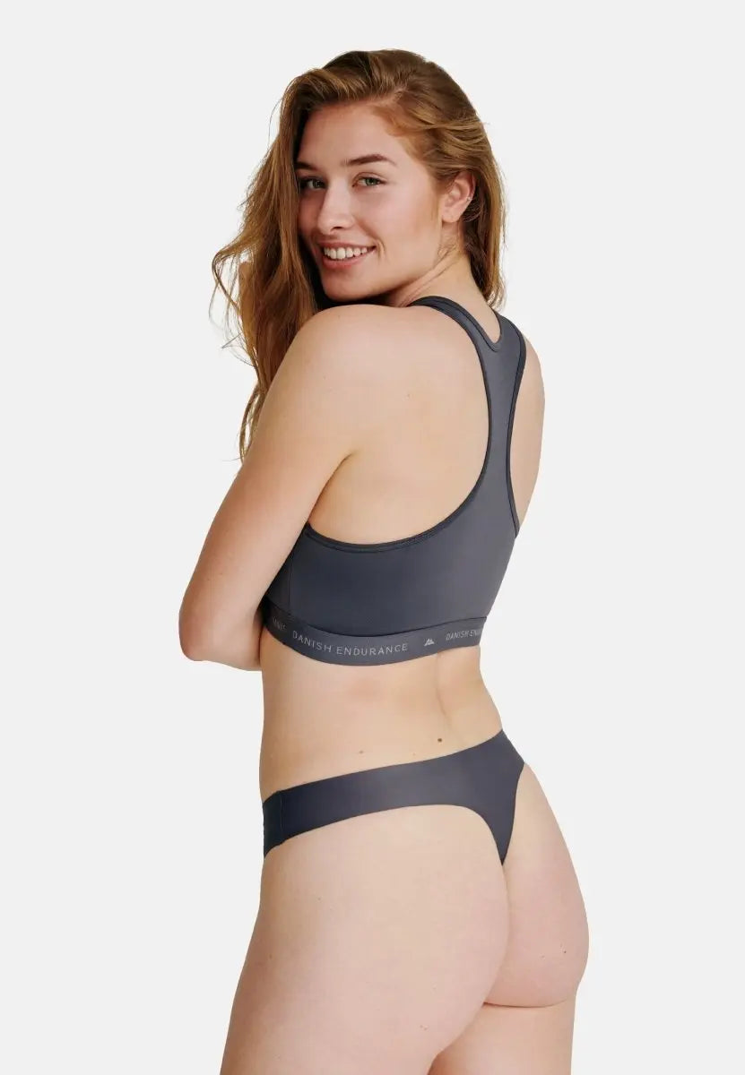 Woman in dark gray thong panties and sports bra, smiling over shoulder against white background.