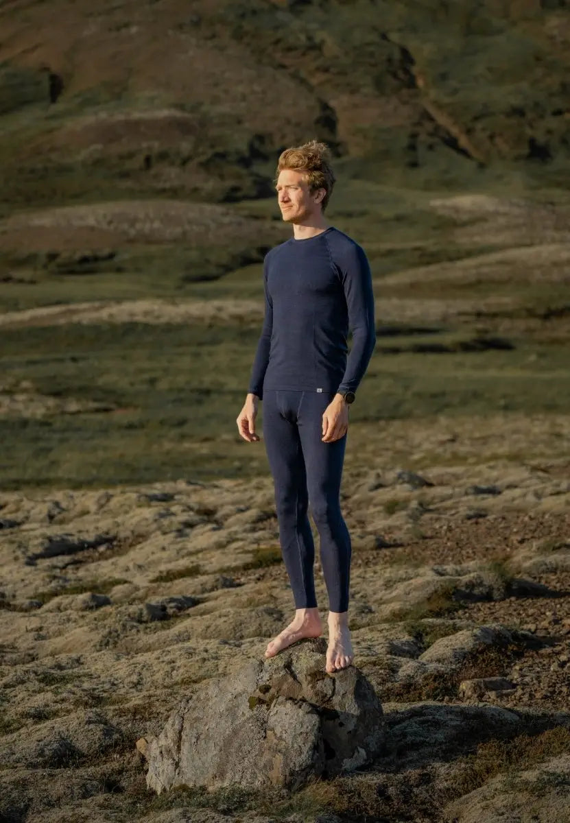 Man in blue merino wool base layer stands barefoot on a rock in a grassy, rocky landscape.