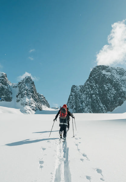 Skier with red backpack trekking through snowy mountains under a clear blue sky.