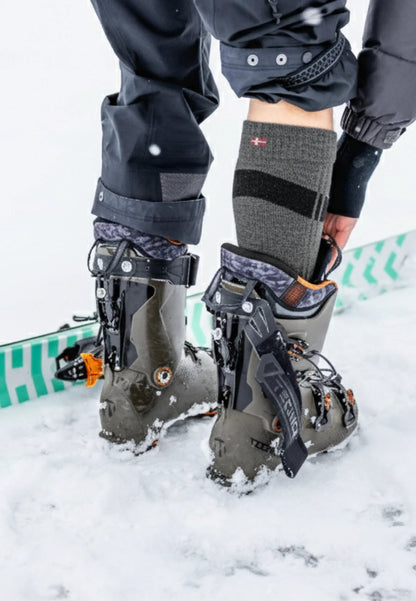 Person adjusting gray Merino wool socks with ski boots in snow, wearing dark pants.